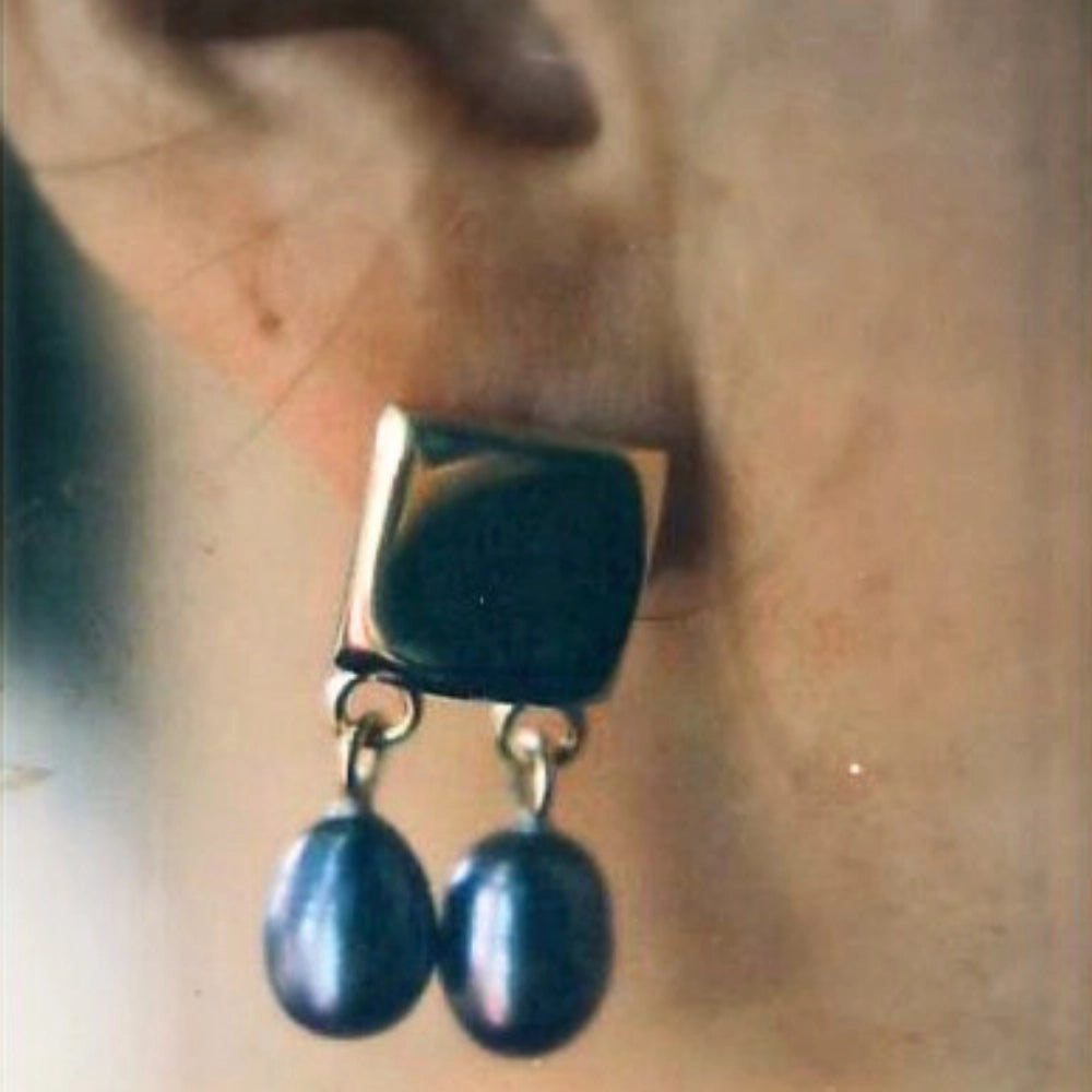 A close-up of a dangling earring in 18ct yellow gold with a square and two hanging oval dark pearl-like drops. Tg-Jewellery